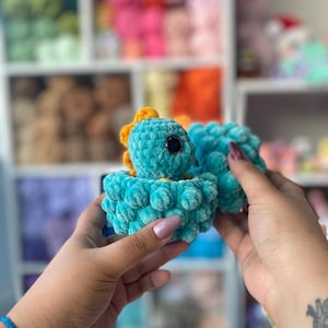 May include: A turquoise crocheted dinosaur toy with a yellow crest and large black eyes. The plush toy is held in a person's hands. The background shows shelves filled with colorful yarn.