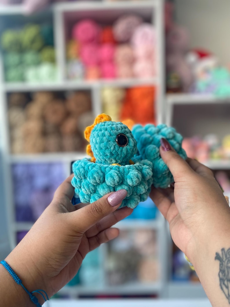 May include: A turquoise crocheted dinosaur toy with a yellow crest and large black eyes. The plush toy is held in a person's hands. The background shows shelves filled with colorful yarn.
