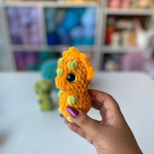 May include: A handmade orange crochet dinosaur with green spots and large black eyes. The plush toy is held in a hand, with other similar toys visible in the background. The dinosaur is a soft, cuddly toy.