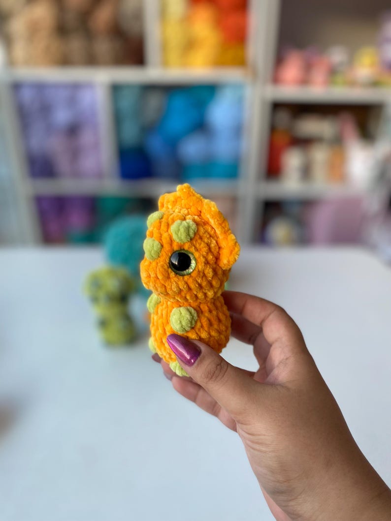 May include: A handmade orange crochet dinosaur with green spots and large black eyes. The plush toy is held in a hand, with other similar toys visible in the background. The dinosaur is a soft, cuddly toy.