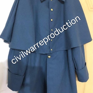 May include: A blue Civil War reproduction coat with a cape-like top layer. The coat has gold-colored buttons and long sleeves with folded cuffs. The text "civilwarreproduction" is printed on the coat.