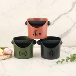 May include: Three personalized coffee tampers in different colors: coral, olive green, and black. Each tamper has a black handle and a name or initial engraved on the front. The coral tamper is labeled "Ava", the green one "Kate", and the black one "Stuart".