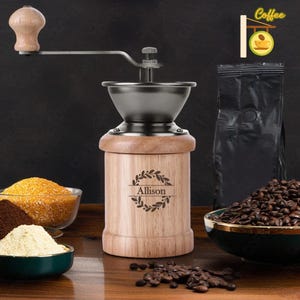 Peut inclure: Un moulin à café manuel en bois avec une manivelle en métal et une trémie en métal. Le moulin porte une inscription personnalisée qui dit "Allison" entourée d'une couronne. Le moulin est posé sur une table en bois avec des grains de café et d'autres ingrédients.
