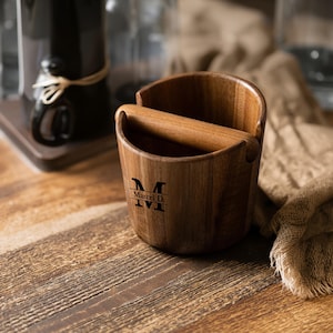 Peut inclure: Un récipient en bois pour vider le marc de café, de forme incurvée, avec une barre en bois. Le récipient porte l'inscription "M Marcel D. IVI" sur le devant. Il est posé sur une surface en bois.