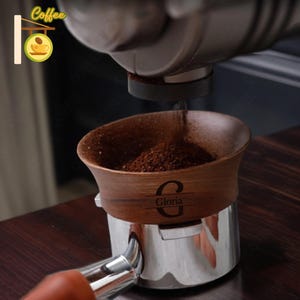 May include: A close-up of a wooden portafilter with a personalized inscription "Gloria" and a dark brown coffee grounds inside. The portafilter is being filled with coffee grounds from a grinder.