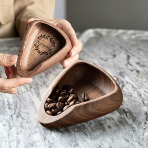 May include: Two wooden, triangular-shaped bowls. One bowl is filled with coffee beans. The other bowl has the name "Natassha" engraved inside, surrounded by a laurel wreath design. The bowls are held against a marble surface.