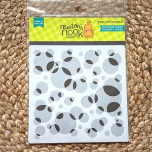 May include: A clear plastic package containing a white stencil with a pattern of overlapping circles, some with dark gray accents. The stencil is labeled "purr-fect stencil" and "multi-layer design includes 2 pieces". The package reads "made in the USA".