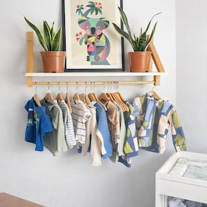 May include: A wall-mounted wooden shelf with a hanging rail displaying various children's shirts and jackets on wooden hangers. Two potted snake plants sit on the shelf beside a framed koala print. A white changing table is visible below.
