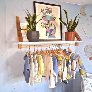 May include: A white shelf with a clothes rail below, displaying children's garments. Above, a framed koala print and potted plants. The light blue wall features decorative painted elements.