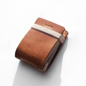 以下が含まれることがあります： ベージュのゴムバンドが付いた茶色の木製ノート。表紙には「leather」の文字がエンボス加工されています。ノートは閉じられており、複数のページで構成されているようです。背景は白です。