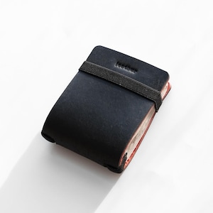Op de afbeelding: Een zwart notitieboek met een zwarte elastische band sluiting. Het woord "leather" is in de bovenste omslag geëmbosseerd. De pagina's hebben een roodbruine tint. Het notitieboek staat op een witte achtergrond.