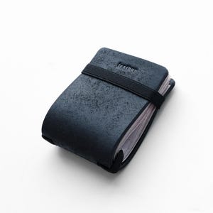 Op de afbeelding: Een donkerblauwe kaarthouder met een getextureerd oppervlak en een zwarte elastische band. Het woord "leather" is in de bovenkant geëmbosseerd. De kaarthouder staat op een witte achtergrond.