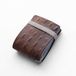 Può includere: Un portafoglio in pelle marrone con una trama simile alla pelle di coccodrillo. Chiuso da un elastico grigio. Design rettangolare e classico, adatto all'uso quotidiano.