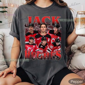 May include: Dark gray t-shirt featuring a graphic of Jack Hughes and other hockey players in red and black jerseys. The name "JACK HUGHES" is printed in red, with a vintage style design.