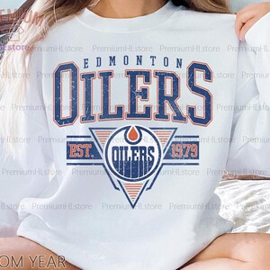 Puede incluir: Sudadera blanca con el texto "EDMONTON OILERS" en azul y naranja, con un efecto vintage desgastado. El diseño incluye el logotipo del equipo y el año 1979. Una prenda superior informal de manga larga.