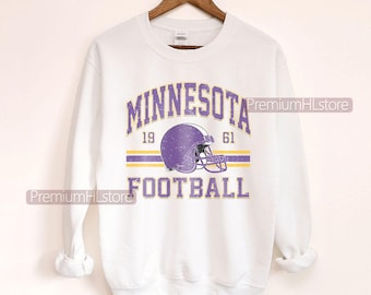 Puede incluir: Sudadera blanca con la inscripción "MINNESOTA FOOTBALL" en morado, y un gráfico de casco de fútbol morado. El diseño incluye los años 1961 y 1961, y dos rayas amarillas. Las palabras "CUSTOM YEAR" están en la parte inferior.