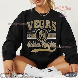 Puede incluir: Sudadera negra con texto dorado "VEGAS Golden Knights EST. 2016". El dise&ntilde;o incluye un emblema circular con un casco de caballero. Tambi&eacute;n son visibles las palabras "CUSTOM YEAR" y "BLACK".