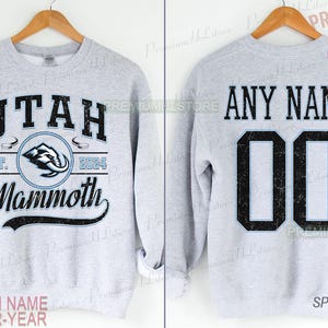K&ouml;nnte beinhalten: Hellgraues Sweatshirt mit der Aufschrift "UTAH Mammoth" und einem Mammut-Logo auf der Vorderseite. Auf der R&uuml;ckseite steht "ANY NAME" und die Nummer "00". Das Sweatshirt ist sportgrau.