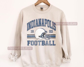 May include: A beige sweatshirt featuring the text "INDIANAPOLIS FOOTBALL" in blue, and a football helmet graphic. The sweatshirt has a crew neck and long sleeves. The text "1953" is also visible.