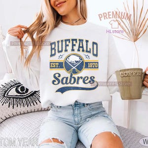 May include: White sweatshirt featuring the text "BUFFALO SABRES" in blue and gold, with a logo and the year "1970". The design has a vintage, distressed look. The sweatshirt is a crew neck style.