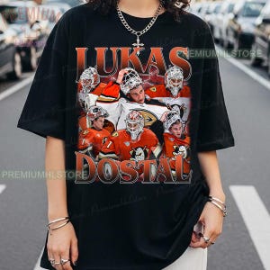 May include: Black t-shirt featuring a collage of a hockey goalie in action, with the name "LUKAS DOSTAL" in orange lettering. The goalie is wearing a helmet and jersey with the number 1. The shirt has a vintage, distressed look.