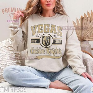 Puede incluir: Sudadera de color arena con texto y logotipo de "VEGAS Golden Knights". El dise&ntilde;o incluye el nombre del equipo, un logotipo de escudo y el a&ntilde;o "2016". La sudadera la lleva una persona sentada.