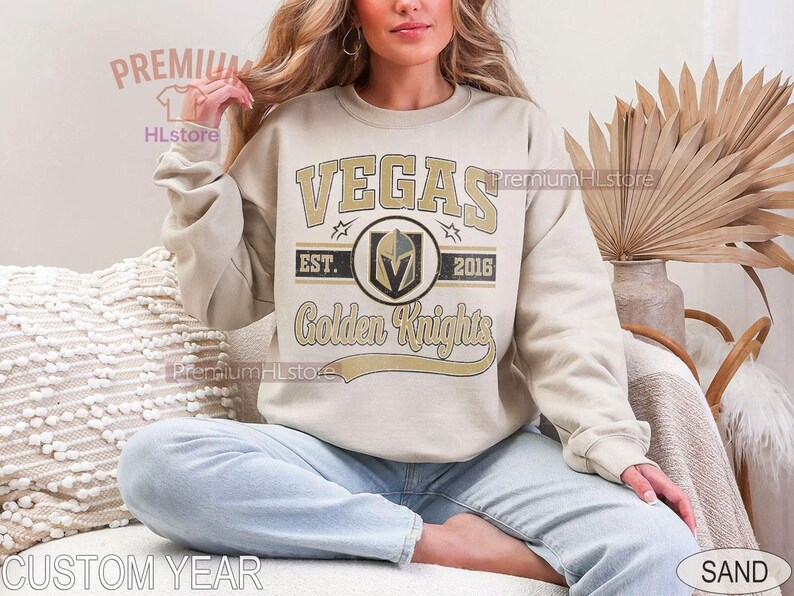 Puede incluir: Sudadera de color arena con texto y logotipo de "VEGAS Golden Knights". El dise&ntilde;o incluye el nombre del equipo, un logotipo de escudo y el a&ntilde;o "2016". La sudadera la lleva una persona sentada.