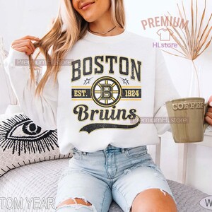 Puede incluir: Sudadera blanca con el texto "BOSTON Bruins EST. 1924" en negro y dorado. El dise&ntilde;o incluye un logotipo de los Bruins. La sudadera es de estilo casual, adecuada para los fan&aacute;ticos del deporte.