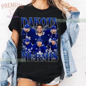 May include: Black t-shirt featuring a graphic of hockey players in blue uniforms. The text "DAKOTA MERMIS" is displayed in a stylized font above and below the players. The shirt has a crew neck and short sleeves.