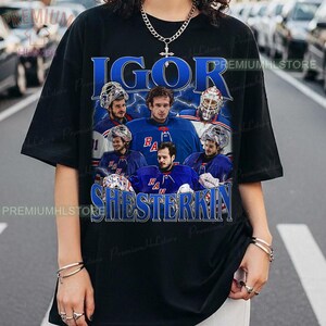 Puede incluir: Camiseta negra con un diseño gráfico azul del jugador de hockey Igor Shesterkin. El diseño incluye los nombres "IGOR" y "SHESTERKIN" en azul, junto con imágenes del jugador con su uniforme de hockey.