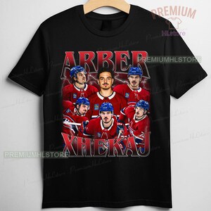 May include: Black t-shirt featuring a graphic of a hockey team with the names "ARBER" and "XHEKAJ" in red. The team is wearing red and blue uniforms. The shirt has a vintage, distressed look.