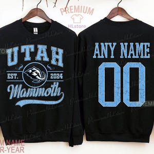 K&ouml;nnte beinhalten: Schwarzes Sweatshirt mit hellblauem Text und Grafiken. Die Vorderseite zeigt "UTAH", "EST. 2024" und "Mammoth" mit einem Logo. Auf der R&uuml;ckseite steht "ANY NAME" &uuml;ber der Nummer "00".