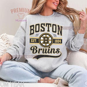 Puede incluir: Una sudadera gris claro con el logotipo de los Boston Bruins en negro, amarillo y blanco. El dise&ntilde;o incluye el nombre del equipo, un logotipo circular con una 'B' y el texto 'EST. 1924'. La sudadera es de estilo casual.