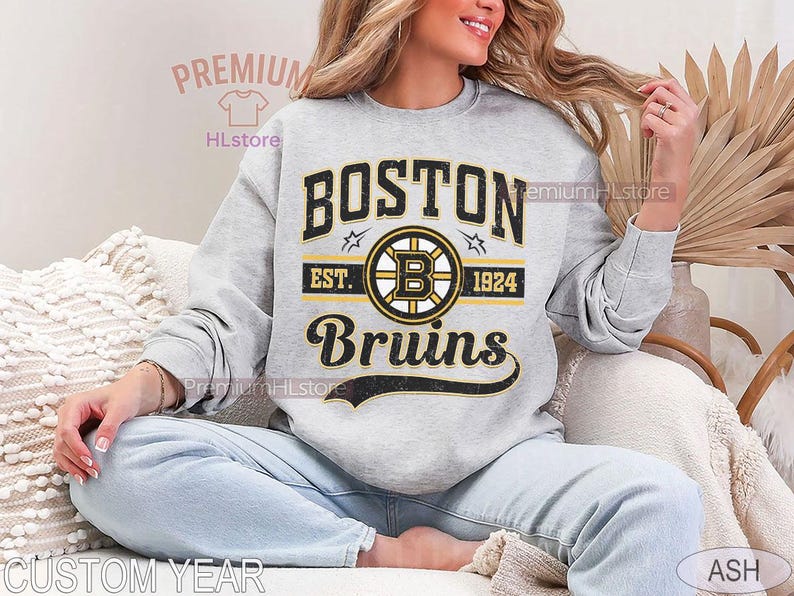 Puede incluir: Una sudadera gris claro con el logotipo de los Boston Bruins en negro, amarillo y blanco. El dise&ntilde;o incluye el nombre del equipo, un logotipo circular con una 'B' y el texto 'EST. 1924'. La sudadera es de estilo casual.