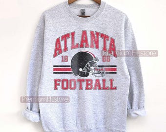 May include: A light grey sweatshirt featuring the text "ATLANTA FOOTBALL" in red, with a black football helmet graphic. The sweatshirt also displays the numbers "19" and "66" in red. The garment is suspended on a wooden hanger.