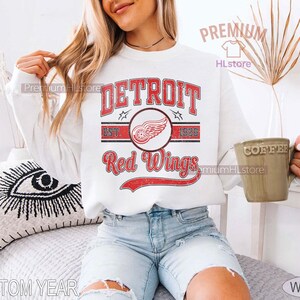 Puede incluir: Sudadera blanca con el texto "DETROIT Red Wings" en rojo, con un logotipo de una rueda alada. El dise&ntilde;o incluye el a&ntilde;o "1926" y una est&eacute;tica vintage. La sudadera es de estilo casual.