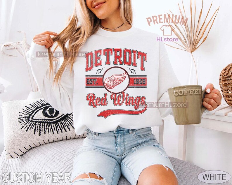 Puede incluir: Sudadera blanca con el texto "DETROIT Red Wings" en rojo, con un logotipo de una rueda alada. El dise&ntilde;o incluye el a&ntilde;o "1926" y una est&eacute;tica vintage. La sudadera es de estilo casual.