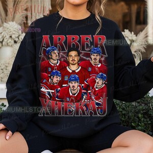 May include: Black sweatshirt featuring a vintage-style graphic of a hockey team with the names "ARBER" and "XHEKAJ" in red and white. The team is wearing red jerseys and blue helmets. The sweatshirt is a crew neck.