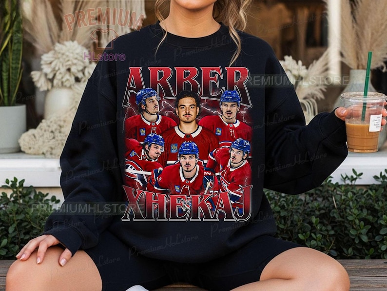 May include: Black sweatshirt featuring a vintage-style graphic of a hockey team with the names "ARBER" and "XHEKAJ" in red and white. The team is wearing red jerseys and blue helmets. The sweatshirt is a crew neck.