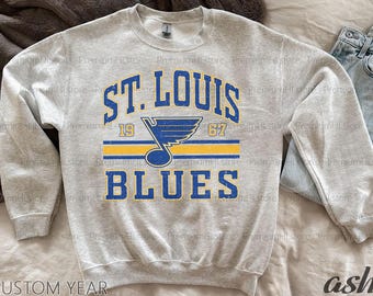 May include: A light gray sweatshirt featuring the text "ST. LOUIS BLUES" in blue lettering with a yellow and blue logo. The numbers "19" and "67" are also visible. The sweatshirt is made of a soft material.
