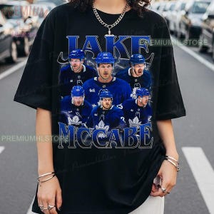 Könnte beinhalten: Schwarzes T-Shirt mit einer Collage des Eishockeyspielers Jake McCabe in blauem Trikot. Der Name des Spielers steht in blauer Schrift oben und unten. Das T-Shirt hat einen Rundhalsausschnitt und kurze Ärmel.