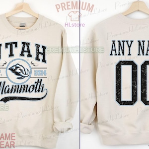 K&ouml;nnte beinhalten: Sandfarbenes Sweatshirt mit dem Text "UTAH Mammoth" in Schwarz und Hellblau sowie einer Mammut-Grafik. Auf der R&uuml;ckseite steht "ANY NAME" &uuml;ber der Nummer "00".