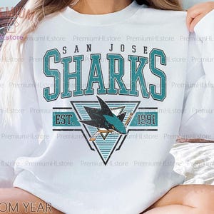 May include: White sweatshirt featuring the San Jose Sharks logo in teal and black. The design includes the team name, a shark graphic, and the year 1991. The sweatshirt has a crew neck and long sleeves.