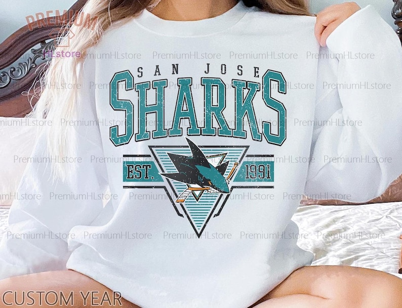 May include: White sweatshirt featuring the San Jose Sharks logo in teal and black. The design includes the team name, a shark graphic, and the year 1991. The sweatshirt has a crew neck and long sleeves.