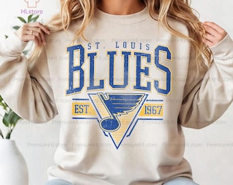 Può includere: Una felpa color sabbia con la scritta "ST. LOUIS BLUES" in blu e oro, un logo e l'anno "1967". La felpa ha maniche lunghe e girocollo. Il marchio "HLstore" è visibile in alto a sinistra.