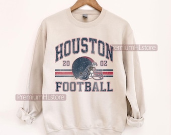 Op de afbeelding: Een beige sweatshirt met de tekst "HOUSTON FOOTBALL" in marineblauw, rood en wit. Het ontwerp bevat een footballhelm en de cijfers "20 02". De sweatshirt hangt aan een houten hanger.