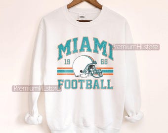 Könnte beinhalten: Weißes Sweatshirt mit einem verblassten türkis-orangenen Aufdruck. Der Aufdruck lautet "MIAMI FOOTBALL" mit einem Football-Helm und den Jahreszahlen "1966" und "19". Das Sweatshirt hängt an einem Holzkleiderbügel.