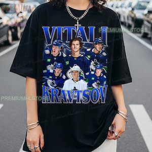 May include: Black t-shirt featuring a graphic of a hockey player and the name "VITALI KRAVTSOV" in blue. The design includes images of the player in a hockey uniform. The shirt has a crew neck and short sleeves.