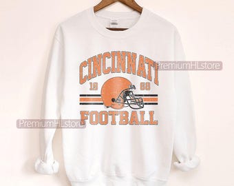 Può includere: Felpa bianca con grafica arancione e nera sbiadita. La grafica recita "CINCINNATI FOOTBALL" con un casco da football e i numeri "19" e "68". La felpa è appesa a una gruccia di legno.