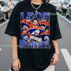 Könnte beinhalten: Schwarzes T-Shirt mit einer Grafik von Eishockeyspielern in Blau und Orange. Der Text "LEON DRAISAITL" ist blau. Das T-Shirt hat einen Rundhalsausschnitt und kurze Ärmel.